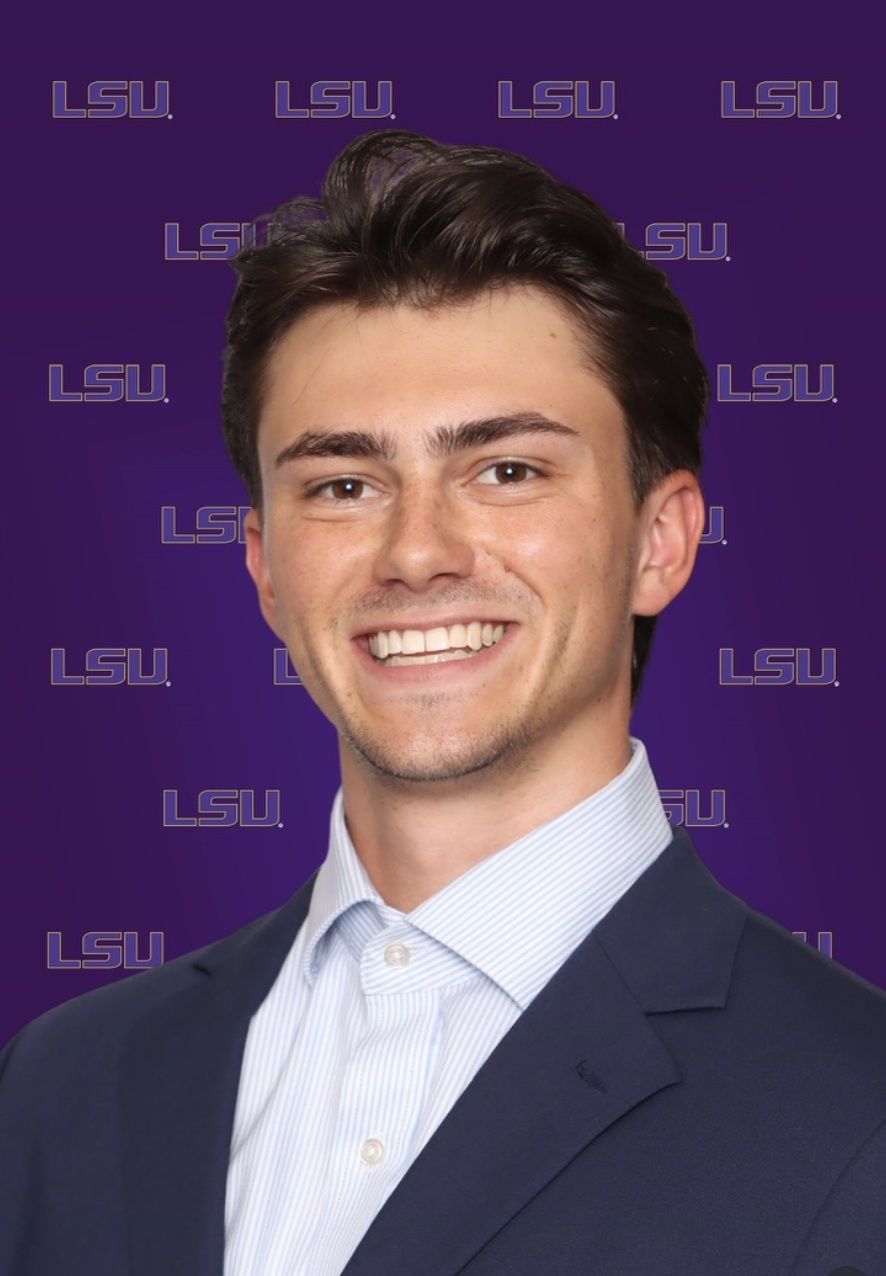A head-and-shoulders portrait of a person wearing a dark suit jacket and a light-colored collared shirt. The background is purple and patterned with repeated “LSU” text in gold lettering. The person is facing forward against the branded backdrop.