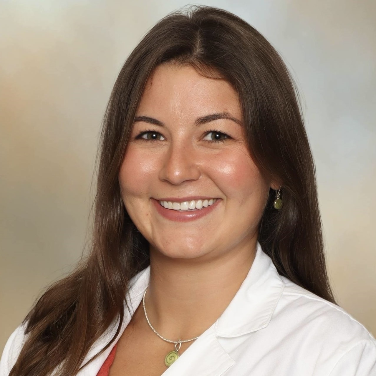 Head-and-shoulders studio portrait of a person with long dark hair wearing a white medical-style coat over a rust-colored top, facing the camera against a softly blended beige and gray background. A necklace and small earrings are visible.