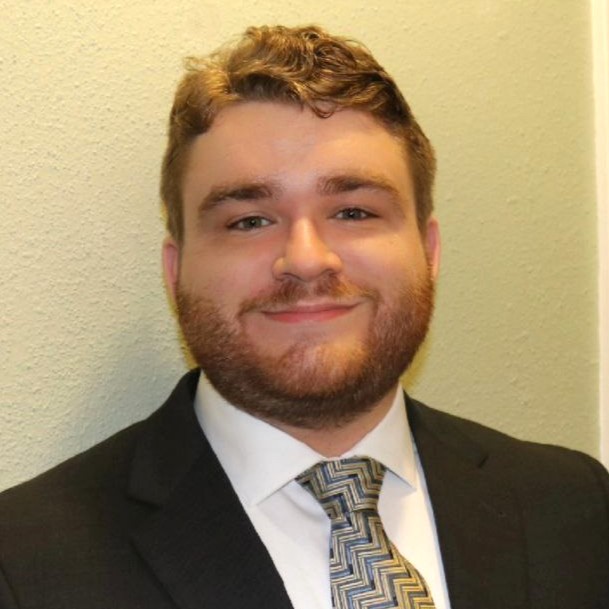 A head-and-shoulders portrait of a person wearing a dark suit jacket, a white dress shirt, and a patterned tie. The person is standing against a light-colored indoor wall with even lighting, and the photo is framed in a formal, professional style.