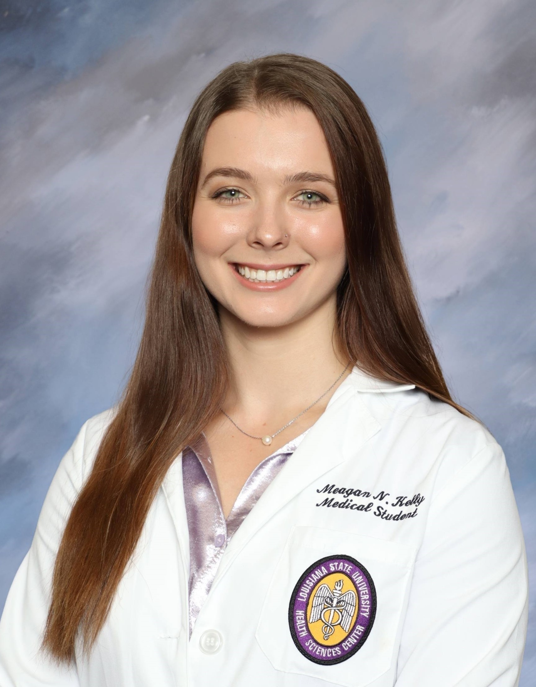 Head-and-shoulders studio portrait of a person with long straight hair wearing a white medical-style coat over a light-colored top, facing the camera against a blue‑gray mottled background. An institutional emblem and embroidered text are visible on the coat.