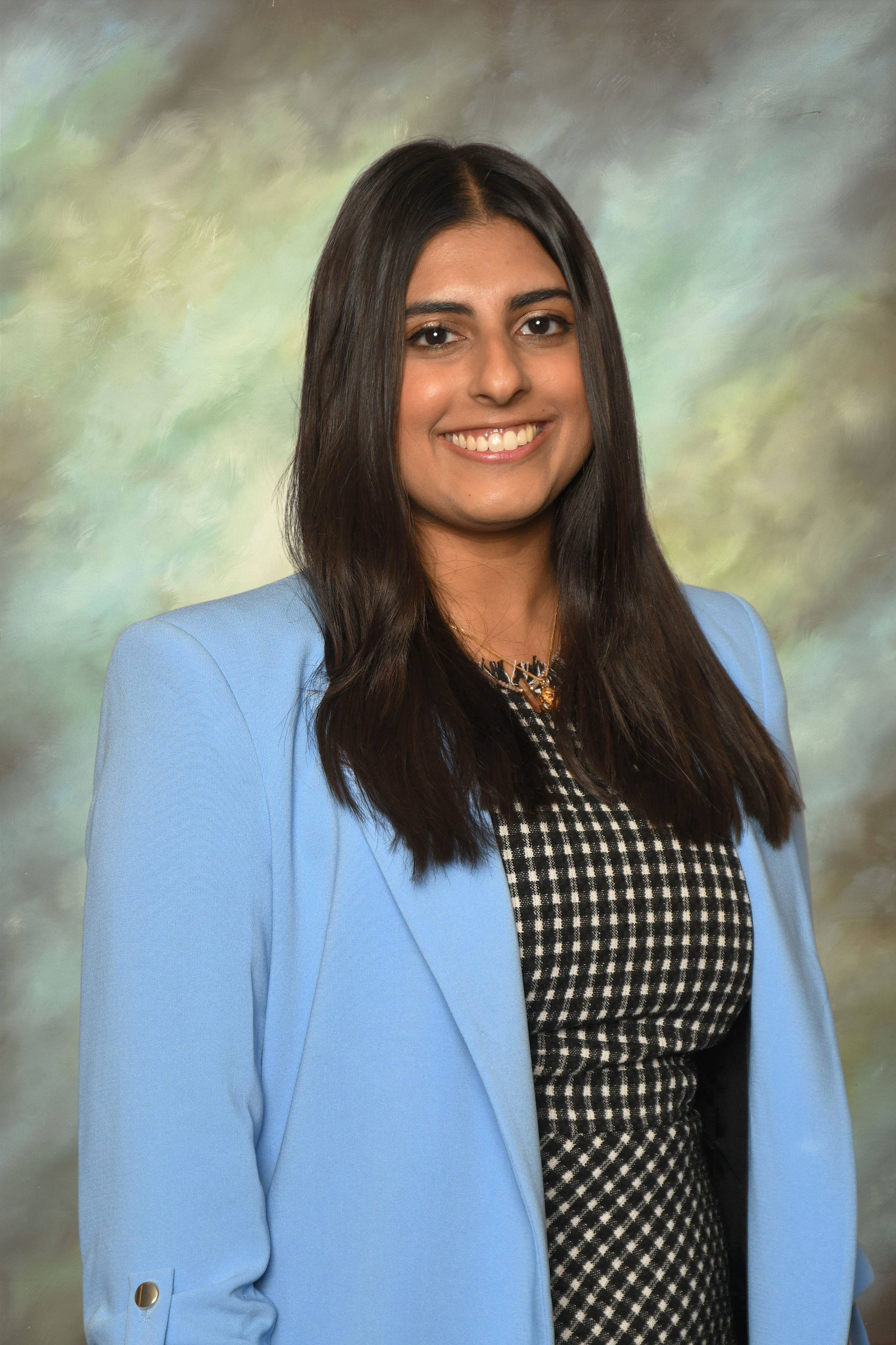 Head-and-shoulders studio portrait of a person with long dark hair wearing a light blue blazer over a black-and-white patterned top, facing the camera against a softly blended green and beige studio background. A necklace is visible.