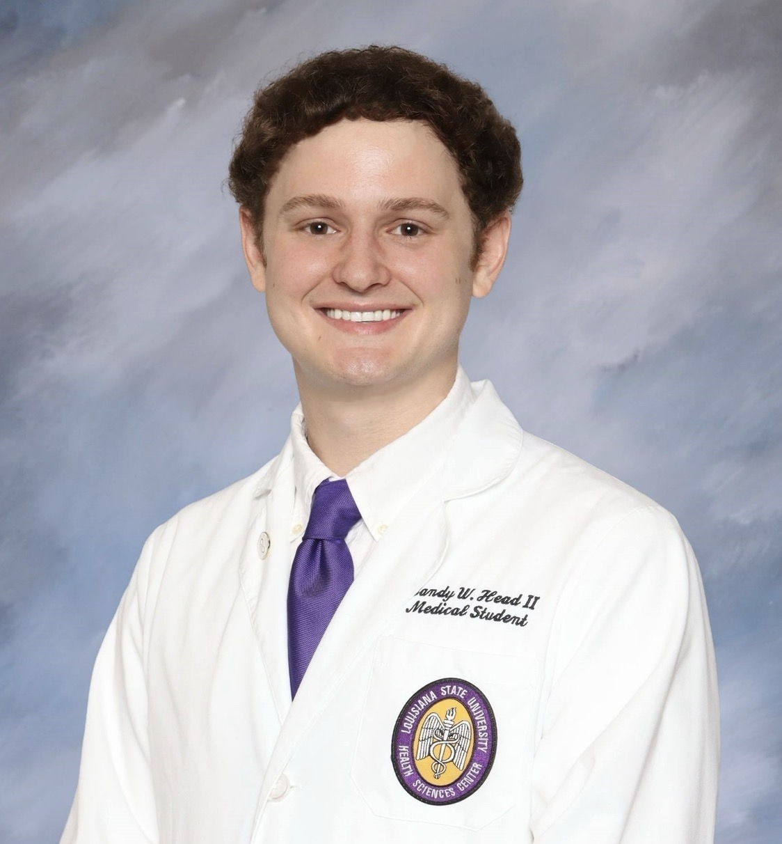 A head-and-shoulders studio portrait of a person wearing a white medical coat and a purple tie. The medical coat features embroidered text and a circular LSU Health Sciences Center patch. The background is a softly blended light blue and gray backdrop, and the subject is facing forward in a professional pose.