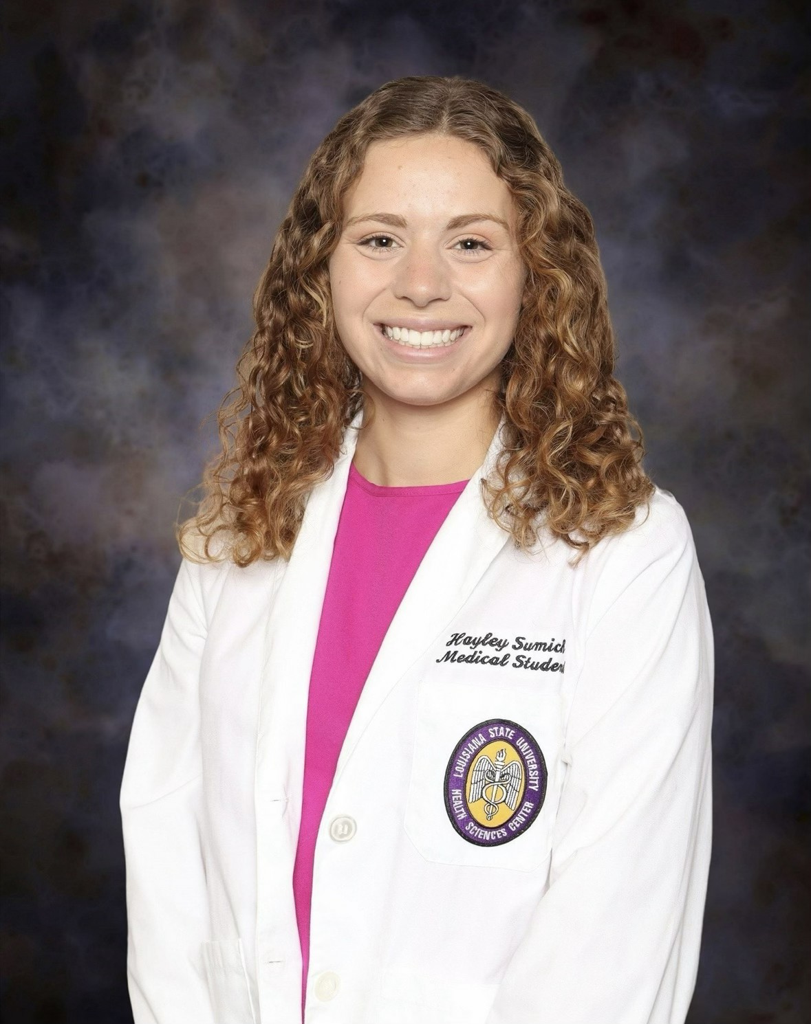 Head-and-shoulders studio portrait of a person with shoulder-length curly hair wearing a white medical-style coat over a bright pink top, facing the camera against a dark, mottled studio background. An institutional emblem and small embroidered text are visible on the coat.