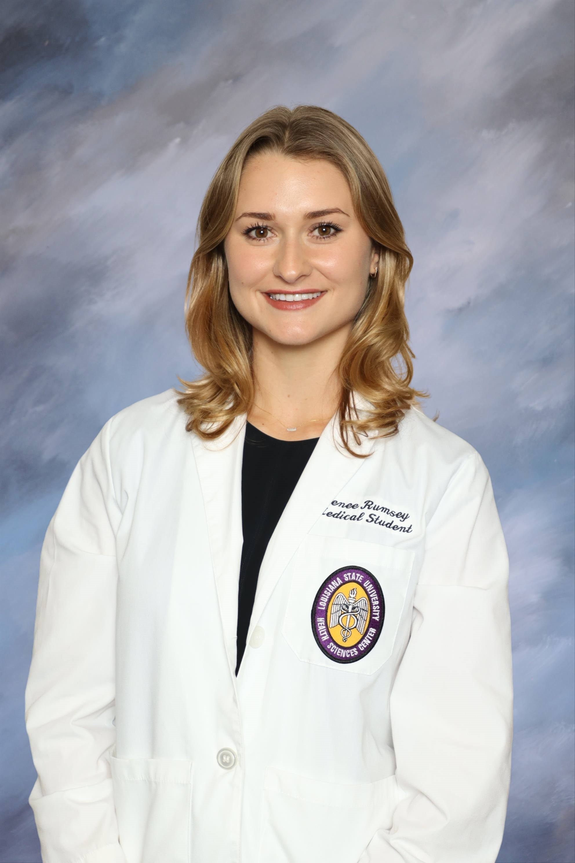 A head-and-shoulders studio portrait of a person wearing a white medical coat over a dark top. The medical coat has embroidered text and a circular LSU Health Sciences Center patch. The background is a softly blended blue and gray studio backdrop, and the subject is facing forward in a professional pose.
