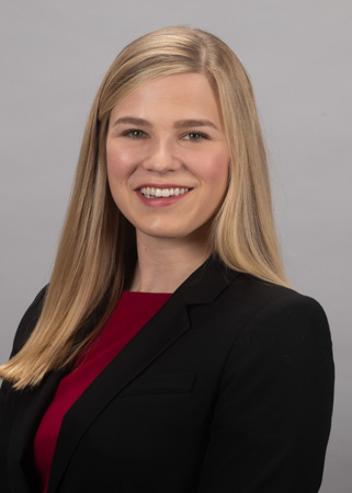 A head‑and‑shoulders studio portrait of a person wearing a black blazer over a red top. The person has long, straight, light‑colored hair worn down over the shoulders. The background is a plain light gray studio backdrop, and the subject is positioned slightly angled while facing toward the camera in a professional pose.