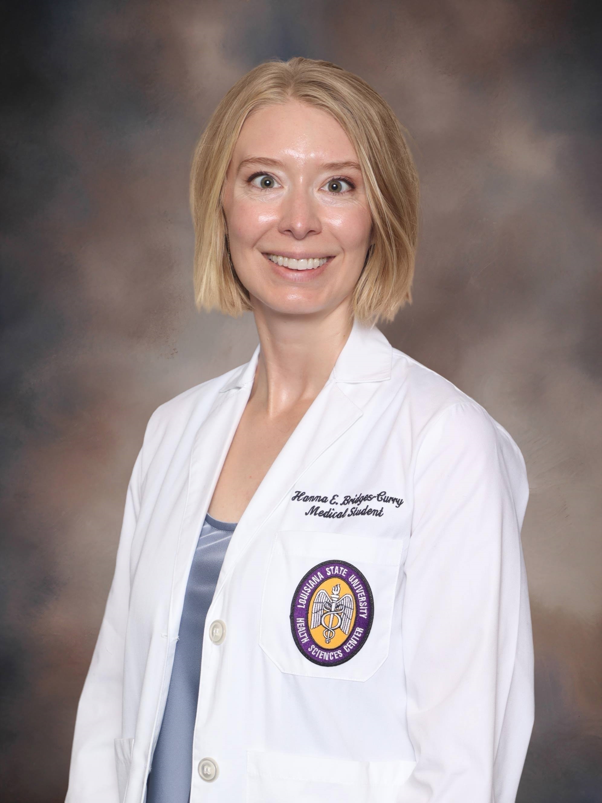 A head-and-shoulders studio portrait of a person wearing a white medical coat over a satin-finish top. The medical coat features embroidered text on the chest and a circular LSU Health Sciences Center patch on the sleeve. The background is a softly blended studio backdrop in muted gray and brown tones, and the subject is facing forward in a professional pose.