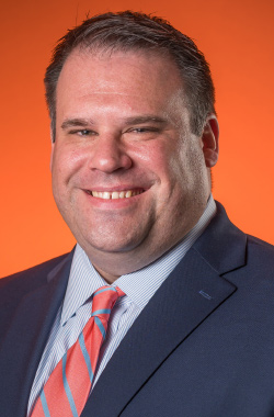 A close-up studio portrait of an adult person wearing a navy blue suit jacket, a light blue striped dress shirt, and a coral-and-light-blue striped tie. The person is facing forward with a slight head tilt and a visible smile. The background is a smooth, solid orange color with even lighting. The image is tightly framed around the head and shoulders, with clear detail on facial features and clothing textures.