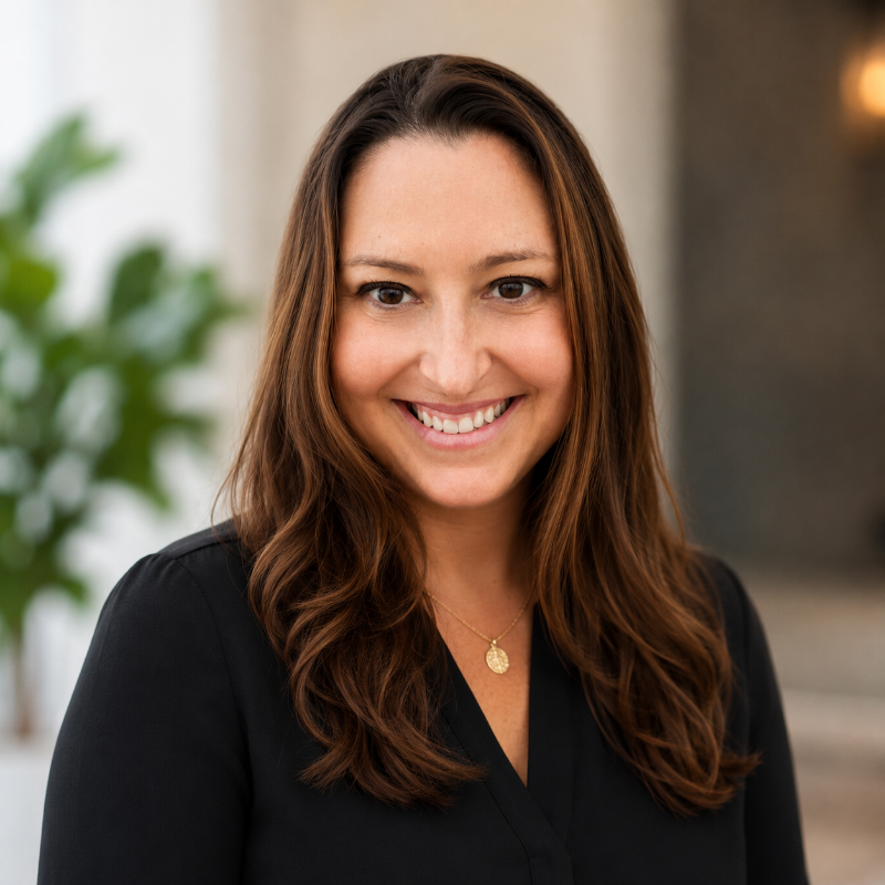 Professional headshot of a person with long brown hair wearing a black blouse and a gold pendant necklace. The person is centered in the frame and looking toward the camera. The background is softly blurred, featuring neutral indoor tones with a green plant visible on the left side and warm lighting on the right.