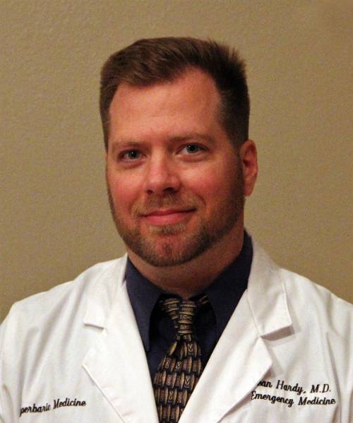 Close-up portrait of an adult individual wearing a white medical coat over a dark shirt and patterned tie, facing slightly to the side against a plain beige background. The coat has embroidered text, including “Emergency Medicine,” visible on the front.