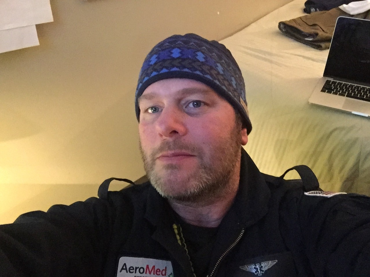 A close-up indoor photo shows a person wearing a dark beanie and a black jacket with an “AeroMed” patch and a winged medical emblem. The person is seated in a room with soft yellow lighting; a bed with folded clothing and an open laptop is visible in the background.