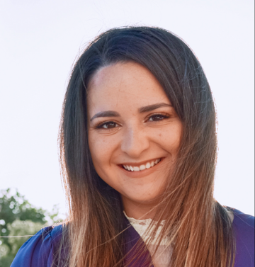 Close-up outdoor portrait of a person with long, straight brown hair parted to one side, wearing a purple top and a light-colored lace neckline. The background is softly blurred with greenery and bright sky, and the lighting is warm and natural.