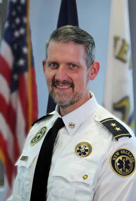 Formal portrait of a uniformed emergency services official in a white dress shirt with shoulder insignia and badge, standing indoors. A circular patch on the sleeve reads “New Orleans Emergency Medical Services,” and a star-of-life emblem is visible on the chest. The background is softly blurred, showing a U.S. flag on one side and a light-colored flag on the other. The person faces forward with a neutral, composed expression under even