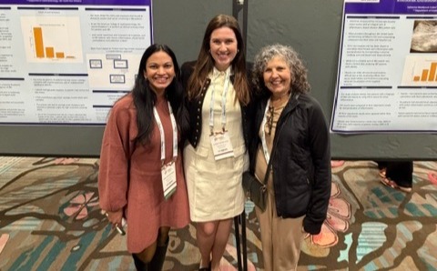 Three conference attendees stand together in front of two LSU Health research poster displays mounted on presentation boards at a professional meeting. The posters feature charts, graphs, text sections, and an ultrasound image, with LSU Health branding visible at the top. The individuals wear conference name badges and business-casual attire, including a light-colored dress, a pink dress with black tights, and a dark jacket with tan pants. The group is posing for a photo in an exhibition hall with patterned carpet and additional poster presentations visible in the background.