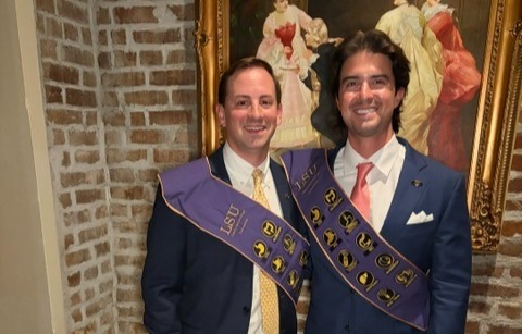 Two adults stand side by side indoors in front of an exposed brick wall and a large framed painting depicting people in historical dress. Both are wearing blue suits, white dress shirts, and ties, along with purple ceremonial sashes decorated with gold lettering and circular emblems. One individual wears a yellow patterned tie, while the other wears a pink tie and white pocket square. The group is facing the camera, and the setting has a formal, celebratory atmosphere with warm lighting and ornate décor.