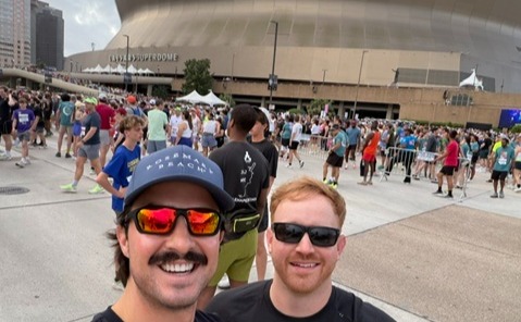 Two adults take a selfie outdoors in front of the Caesars Superdome in New Orleans. The person in the foreground wears a dark baseball cap, reflective sunglasses, and a mustache, while the other wears dark sunglasses and a black shirt. A large crowd is gathered behind metal barricades outside the stadium entrance. The curved exterior of the Superdome dominates the background, with surrounding city buildings visible to the left. The scene appears to be a public event or race, with many attendees wearing athletic clothing and congregating in the plaza area.