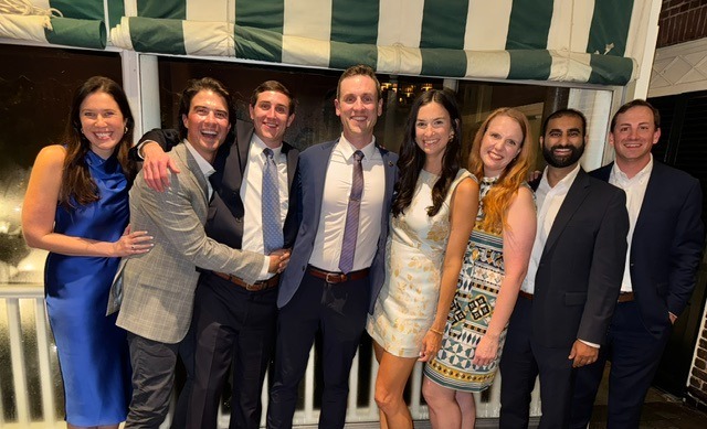 Eight adults pose together on an outdoor patio at night in front of a white railing and green-and-white striped awning. The group is dressed in business and semi-formal attire, including suits, sport coats, dress shirts, and dresses. Several individuals have their arms around one another as they stand closely together facing the camera. Warm indoor lighting is visible through the windows behind them, and the group fills most of the frame in a celebratory gathering setting.