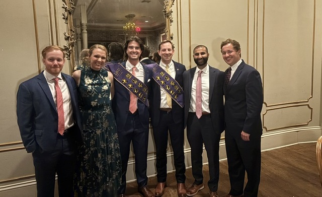 Six adults pose together indoors at a formal celebration, standing in front of an ornate gold-framed mirror and decorative cream-colored wall paneling. The group is dressed in formal attire, including suits, ties, and a patterned floor-length dress. Two individuals in the center wear purple ceremonial sashes with gold lettering and emblems over dark suits. The group stands closely together facing the camera. The room features warm lighting, hardwood floors, and elegant banquet-style décor, creating a festive and celebratory atmosphere.