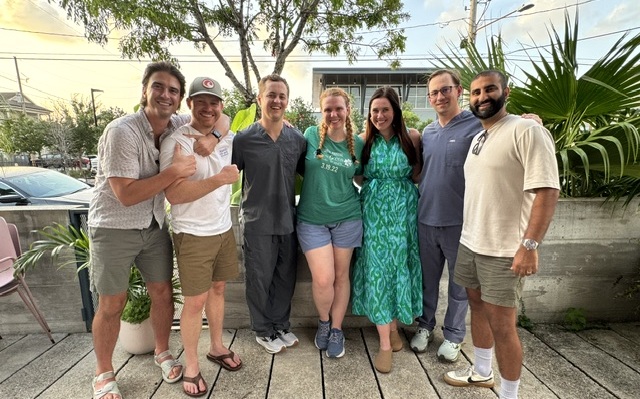 Seven adults stand together outdoors on a patio during daylight, posing for a group photo. The group is dressed in a mix of casual and professional attire, including shorts, a green patterned dress, scrubs, and polo-style shirts. Trees, large leafy plants, parked cars, and a building are visible in the background. The group stands closely together facing the camera, with one person giving a thumbs-up gesture. The setting appears to be a casual social gathering in an urban outdoor space.
