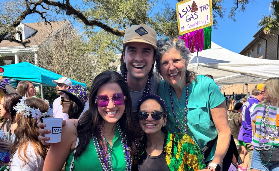 Four people pose together at an outdoor Mardi Gras celebration under a clear blue sky. The group stands in front of festival tents and a crowd, wearing colorful bead necklaces and festive clothing in green, purple, and gold. One person holds a white cup, and another wears a sequined green-and-gold jacket. A handmade sign behind them reads, “LSU Gas Troop me Something!” Trees with sprawling branches frame the scene, and other attendees in Mardi Gras attire fill the background.