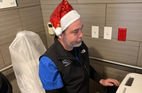 A person wearing blue medical scrubs, a black sleeveless vest with embroidered healthcare-related text, and a red sequined Santa hat sits at a computer workstation in a clinical setting. The person is shown in profile, typing on a keyboard while facing a monitor. The room has gray tiled walls, electrical outlets, and medical equipment visible nearby, including a covered exam chair in the background. The festive holiday hat contrasts with the professional healthcare environment