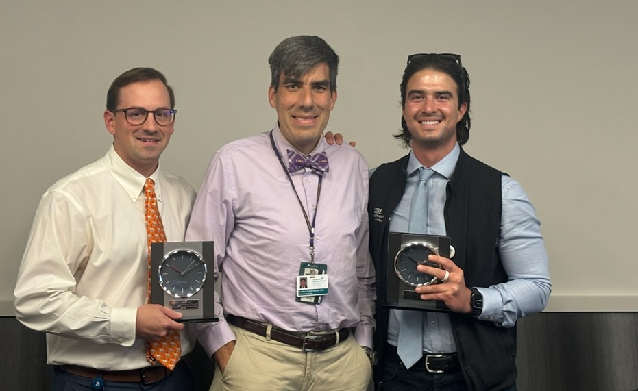 Three people stand together indoors against a plain wall. The two people on the left and right each hold a clock-shaped award plaque, while the person in the center stands between them with an arm around each recipient. Business attire includes dress shirts, ties, and an ID badge worn by the center individual. The awards have dark faces with silver-colored trim and mounted nameplates. The photo appears to have been taken during a recognition or presentation event in a conference or meeting room.