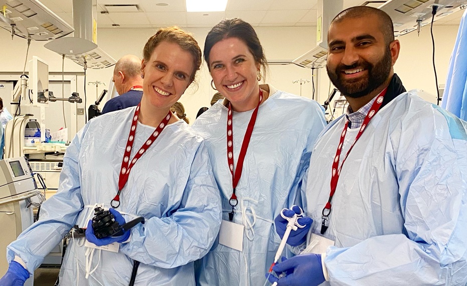 Three individuals wearing light blue disposable medical gowns, blue gloves, and red lanyards stand together in a gastrointestinal endoscopy training or procedure room. One person holds an endoscope control handle, another holds a flexible endoscopic accessory, and the third holds a catheter-like medical device. Medical equipment, monitors, procedure-room ceiling booms, tubing, and packaged supplies are visible throughout the room. The group stands behind a table covered with sterile medical instruments and accessories, with additional clinical staff and equipment visible in the background.