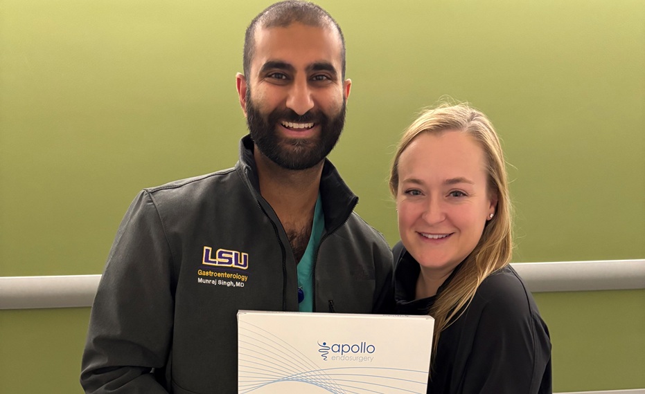 Two individuals stand indoors in front of a light green wall, holding a certificate from Apollo Endosurgery. One individual is wearing a black LSU Gastroenterology jacket with embroidered text, and the other is wearing a black top. Both are facing the camera. The certificate is partially visible and displays the Apollo Endosurgery logo. The setting appears to be a professional or healthcare environment.