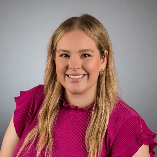 A person with long, light brown hair is shown from the waist up against a plain gray background. They are wearing a bright magenta top with short ruffled sleeves and vertical ribbing. Their posture is relaxed with one arm slightly bent.