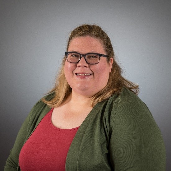 A person with shoulder‑length light brown hair is posed against a plain gray background. They are wearing a dark green cardigan over a red top, sitting slightly angled with relaxed posture.