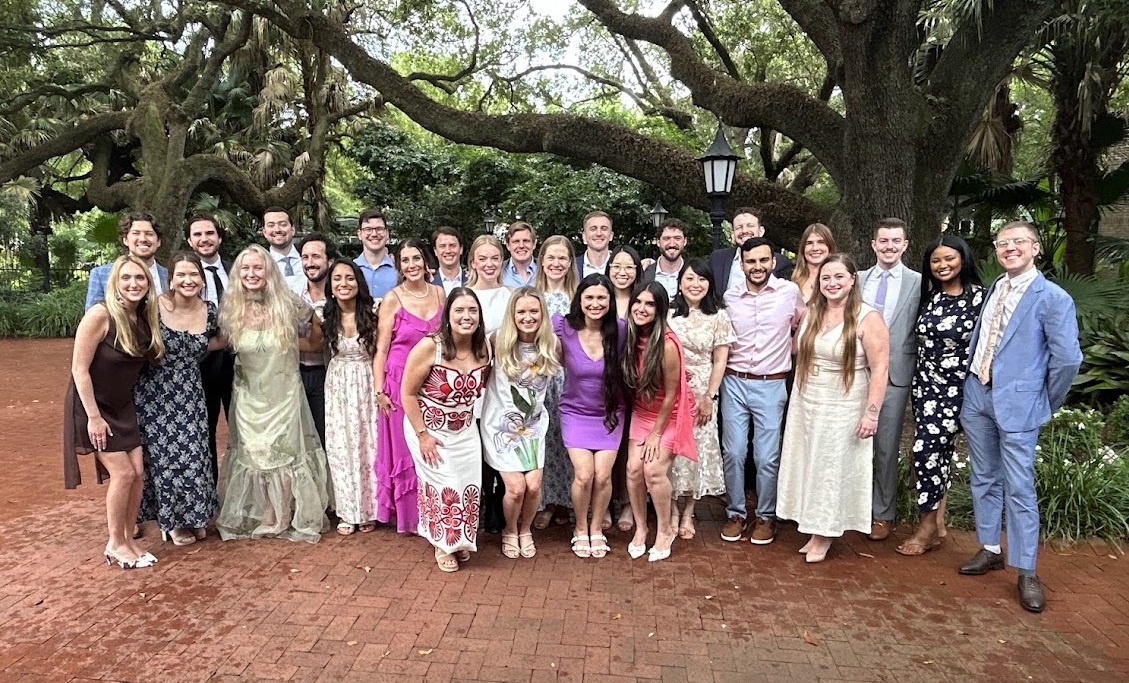 Large group photo of people dressed in formal and semi-formal attire, posing outdoors on a brick walkway beneath large oak trees and surrounded by lush greenery.