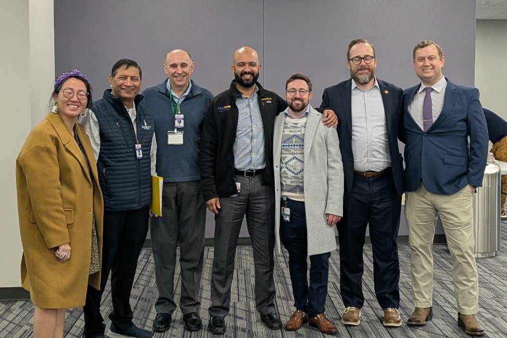 A group of people stand together in a conference or meeting room with gray carpeted floors and neutral-colored walls. They are dressed in a mix of professional and business‑casual attire, including coats, jackets, sweaters, and dress shoes. They stand closely side by side, posing for a group photo.
