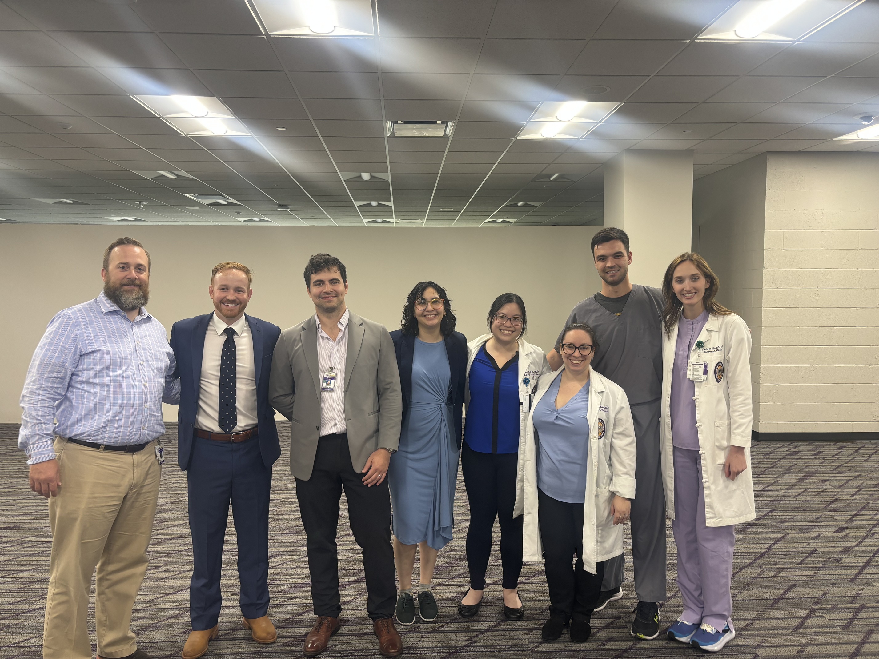 A group of eight medical professionals stand together in a spacious room with carpeted flooring and neutral-colored walls. They are dressed in a mix of business and clinical attire, including dress shirts, suits, scrubs, and white coats. The group stands side by side with arms around one another, posing for a group photo.
