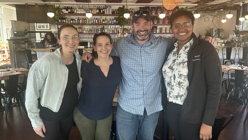 Four people pose together in a restaurant in front of a bar lined with bottles and shelves.