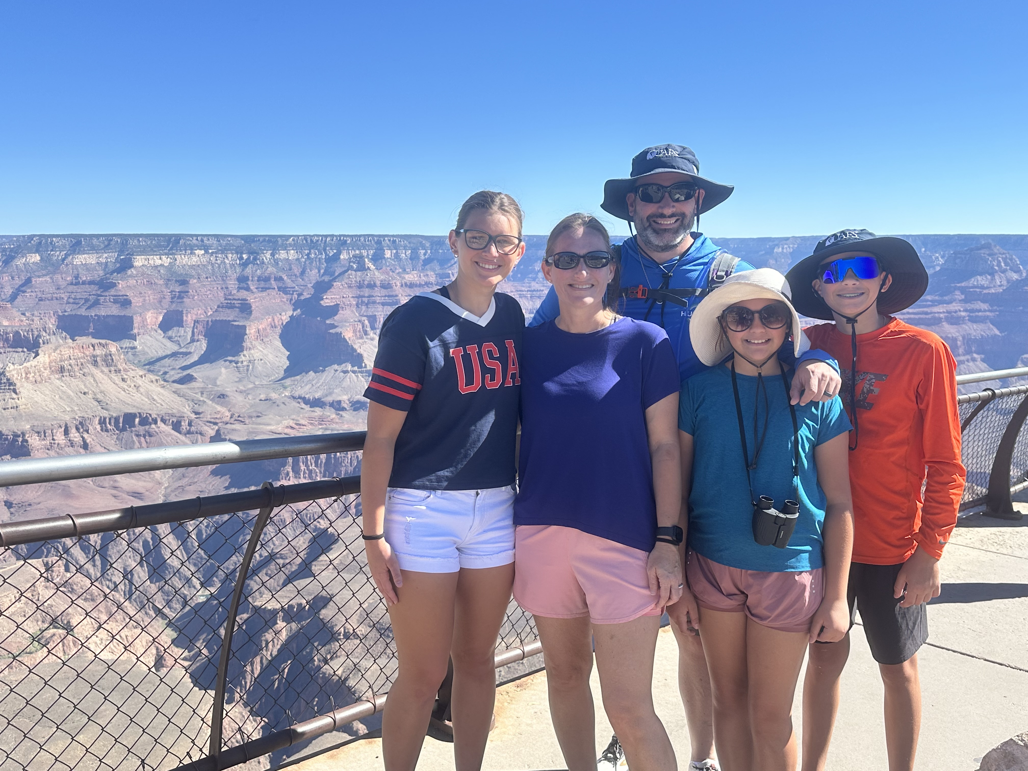Five people pose together at a scenic overlook with the Grand Canyon visible in the background under a clear blue sky. The group stands near a metal safety railing and wears casual outdoor clothing, including sun hats and sunglasses. Distinctive layered rock formations and canyon cliffs stretch across the horizon behind them.