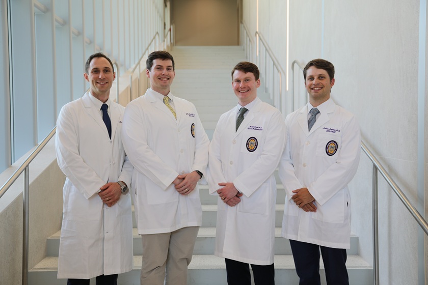 Four people wearing white medical lab coats stand side by side on an indoor staircase in a modern building. The staircase is centered behind the group, with metal handrails and tall windows along one side providing bright natural light. Each person is dressed in professional attire with a collared shirt and tie visible beneath the lab coat. The image is a posed group portrait in a clean, clinical setting.