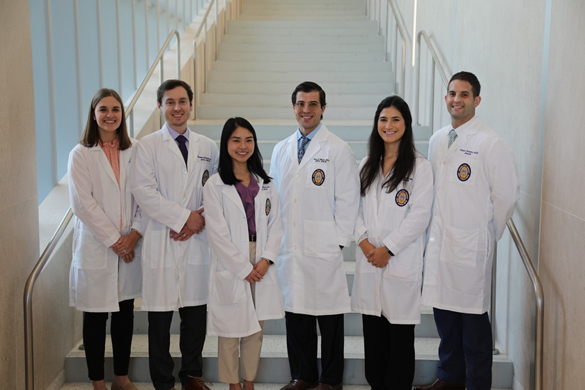 Five people wearing white medical lab coats stand together on a wide staircase inside a bright, modern building. The group is arranged in a single row at the base of the stairs, with several individuals wearing business attire visible beneath the lab coats. Large windows line the left side of the staircase, and metal handrails run along both sides. The white stairway rises behind the group, creating a balanced and professional backdrop with abundant natural light.