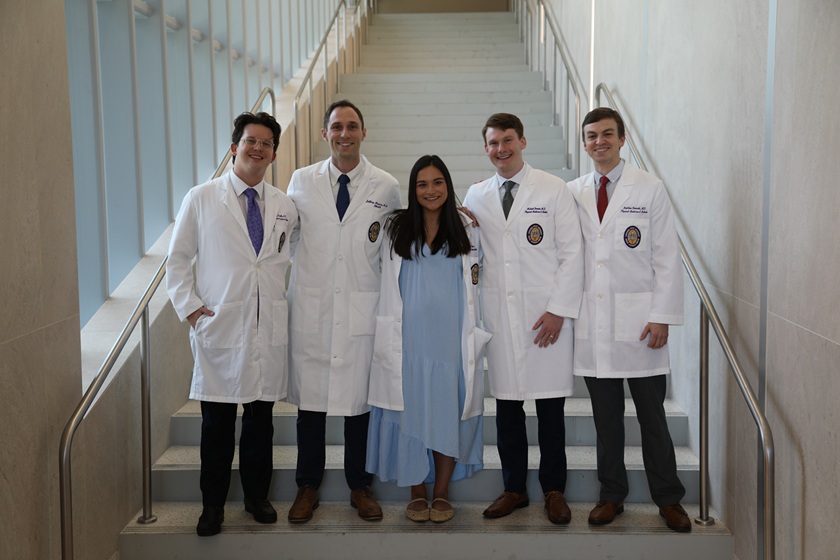 Five individuals pose together on a wide indoor staircase in a modern building with tall windows and metal handrails. Four individuals wear white medical lab coats over professional attire, while the person in the center wears a light blue dress. The group stands in front of a bright stairway that extends upward behind them, creating a symmetrical backdrop. Natural light from the windows illuminates the clean, professional setting.