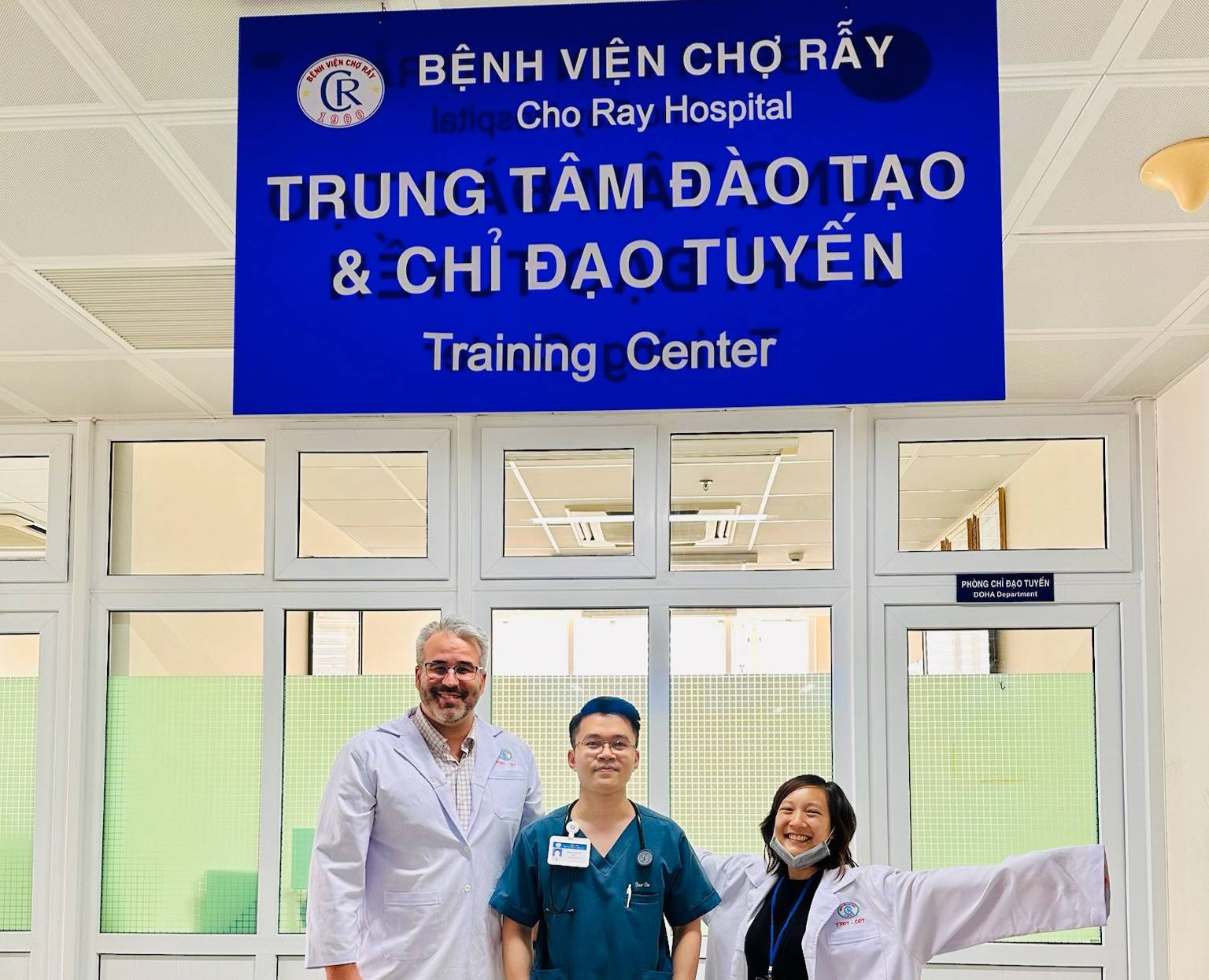 Three individuals wearing medical attire stand together in front of a large blue sign at Cho Ray Hospital. The sign reads ‘Bệnh Viện Chợ Rẫy,’ ‘Cho Ray Hospital,’ and ‘Trung Tâm Đào Tạo & Chỉ Đạo Tuyến,’ with the English translation ‘Training Center’ beneath it. Behind them are white-framed glass doors and a hallway with ceiling lights.
