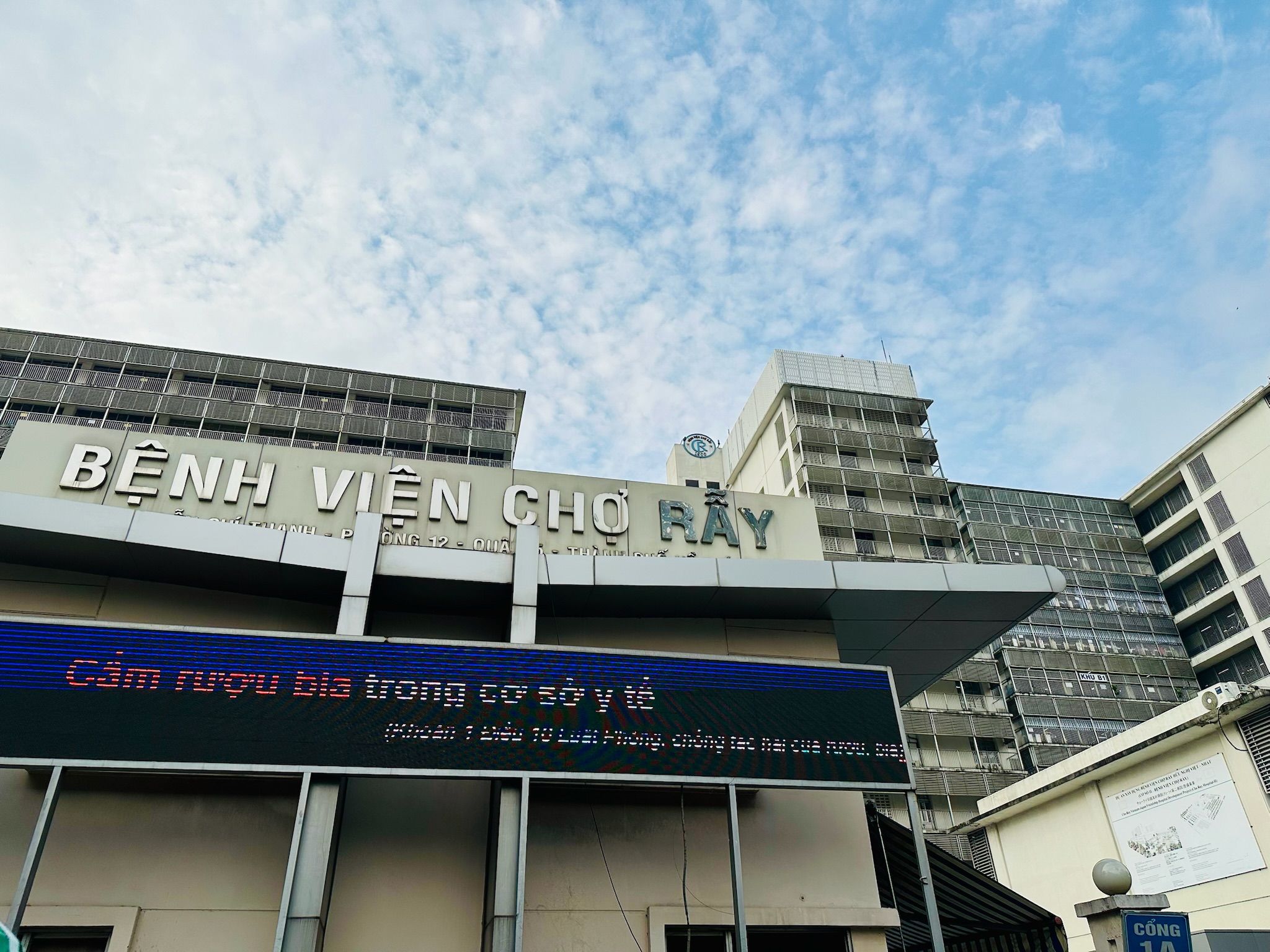 A hospital entrance with a large sign reading ‘Bệnh Viện Chợ Rẫy’ mounted above a modern overhang structure. Below the sign is an electronic display board showing text in Vietnamese. Behind the entrance, a tall multi‑story building with rows of balconies and windows is visible. The sky above is partly cloudy.