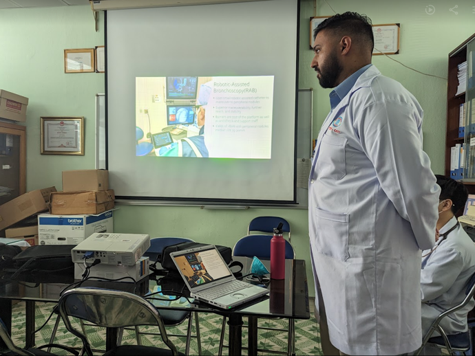 A person wearing a white medical coat stands near a table with a laptop and a projector in a small meeting room. A presentation slide titled ‘Robotic-Assisted Bronchoscopy (RAB)’ is projected onto a screen at the front of the room. Several boxes and framed documents are visible on the wall and shelves in the background, and another person seated at the table is partially visible.