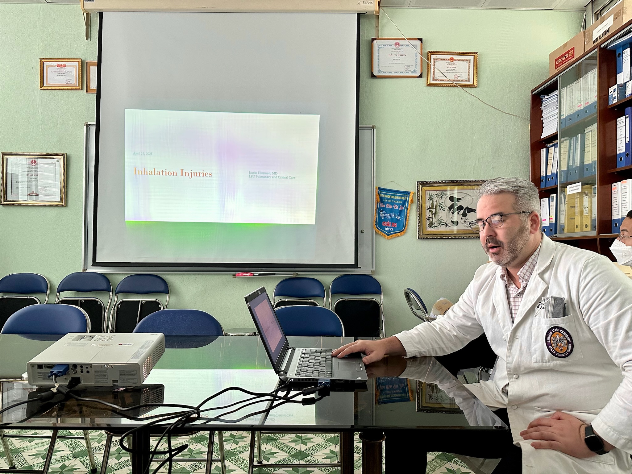 A person wearing a white medical coat sits at a table in a small meeting room, working on a laptop connected to a projector. A presentation slide titled ‘Inhalation Injuries’ is displayed on a projection screen at the front of the room. Several empty chairs are arranged in rows behind the table. Framed certificates and documents are mounted on the wall, and shelves filled with binders are visible on the right side of the room.