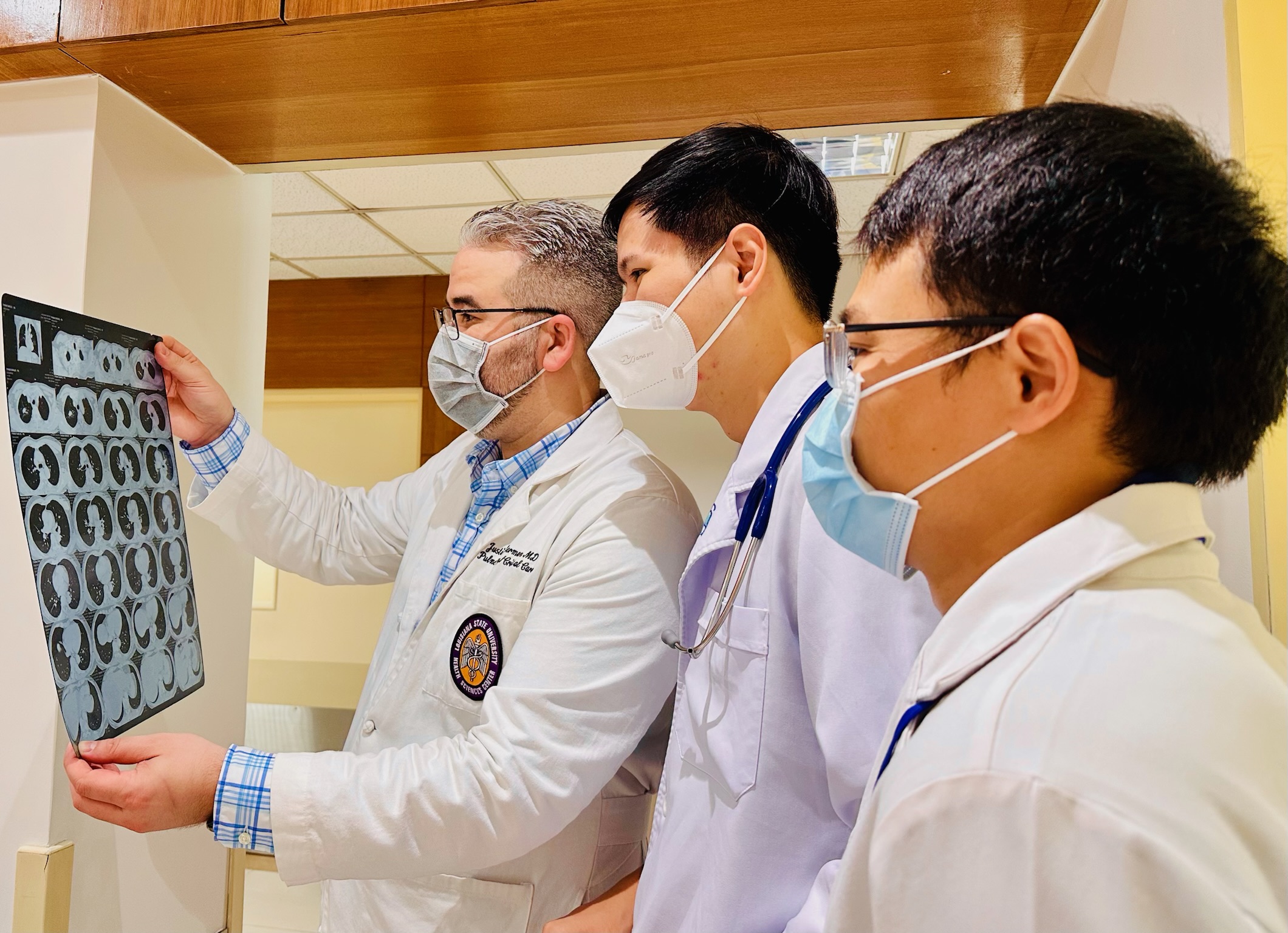 Three people wearing medical coats and face masks stand indoors examining a sheet of medical imaging held up by one of them. The person on the left is holding the film, which contains multiple circular scan images. The two people on the right are looking closely at the film. The setting appears to be a clinical or hospital environment with bright lighting and wood paneling above the doorway.