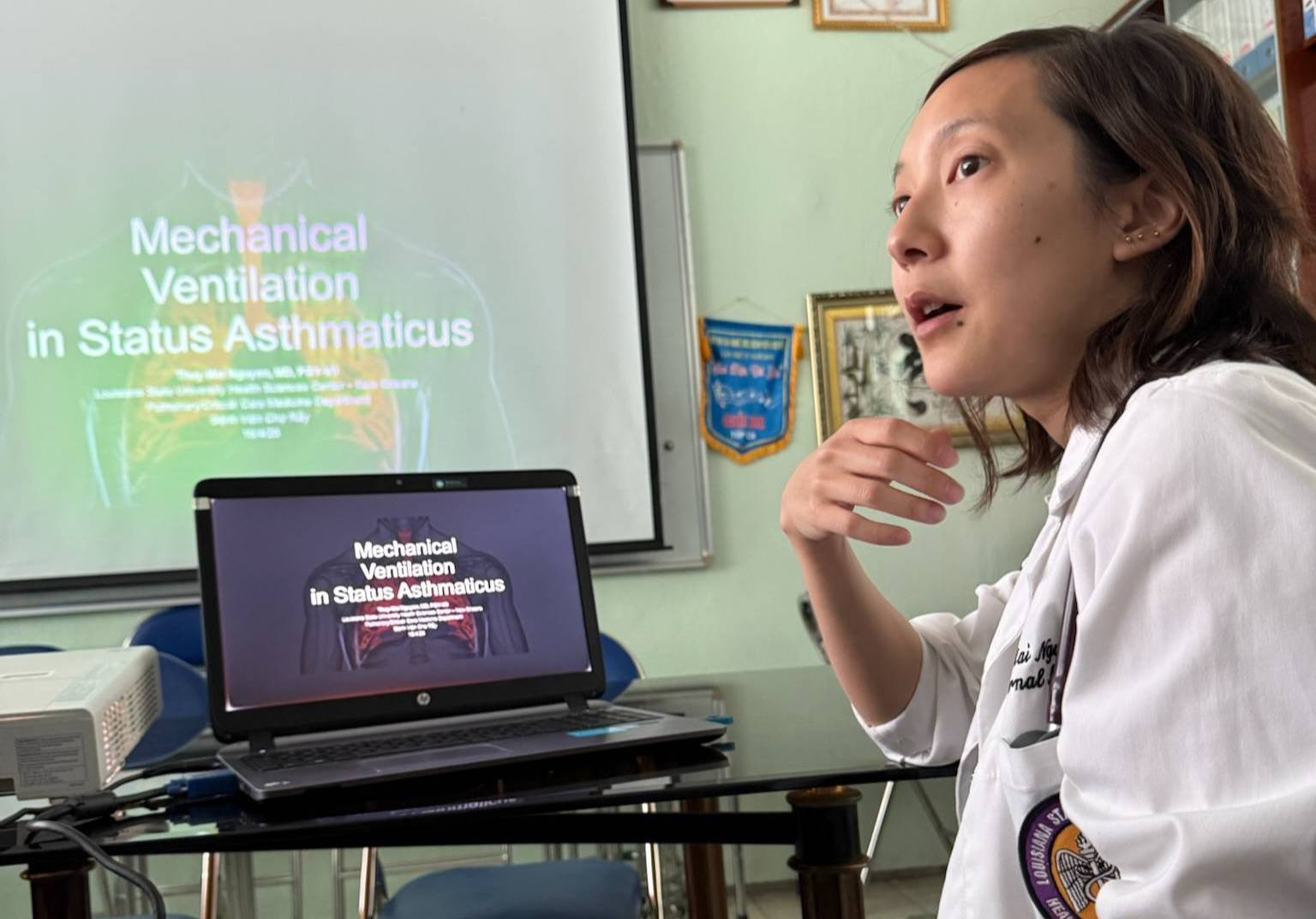 A person wearing a white medical coat sits at a table and appears to be speaking while looking toward the left. On the table is an open laptop displaying a slide titled ‘Mechanical Ventilation in Status Asthmaticus.’ The same slide is projected onto a screen in the background. A projector sits on the table, and framed certificates and banners hang on the wall behind the speaker.