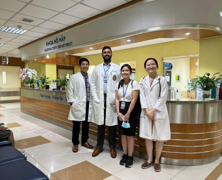 A group of four people in medical attire stand together in front of a hospital reception counter labeled ‘Khoa Hô Hấp’ and ‘Respiratory Department.’ Behind the counter are floral arrangements, office equipment, and signage. The area has tiled floors, overhead lighting, and a curved reception desk with blue Vietnamese text along the front.