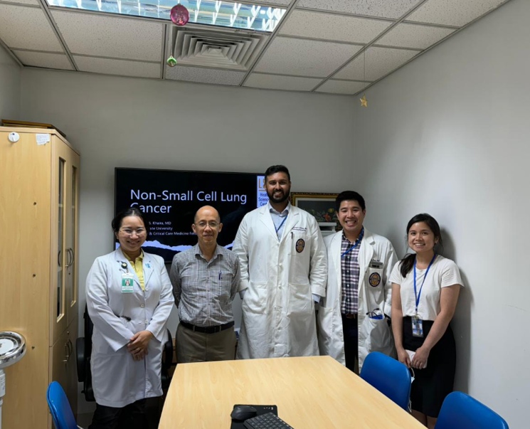 A group of five people stand posing for a photo in a small meeting room. Three individuals are wearing long white coats, one person is wearing a collared shirt, and one person is wearing a short‑sleeved shirt with an ID badge on a lanyard. Behind them, a presentation screen displays the title ‘Non‑Small Cell Lung Cancer.’ A wooden cabinet is on the left side of the room, and a table with blue chairs is in the foreground