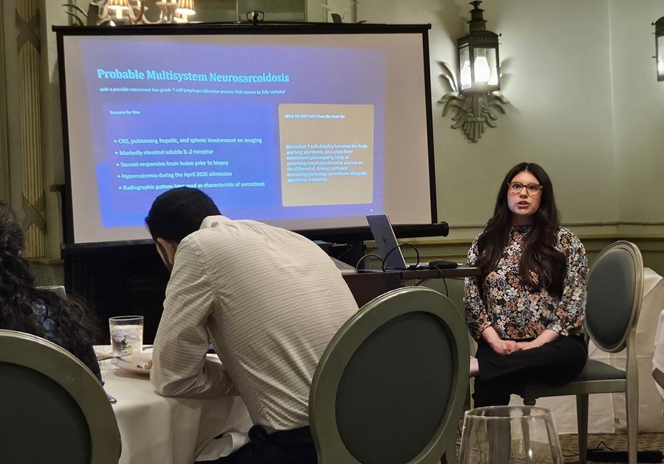 A person seated at the front of a conference room delivers a presentation beside a projection screen displaying a medical slide titled “Probable Multisystem Neurosarcoidosis.” Attendees sit at round tables in the foreground, facing the screen. A laptop is connected to the projector, and wall-mounted lantern-style lights illuminate the room. The setting appears to be a professional meeting or educational presentation in a banquet or conference venue.