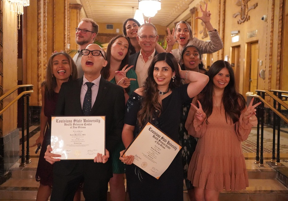 A group of ten individuals pose together in an ornate hallway during a graduation celebration. Two people in the front hold framed diplomas from LSU Health New Orleans while others make playful hand gestures.