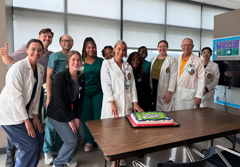 A group of healthcare trainees and faculty members gather around a decorated sheet cake on a table in a clinical setting. Several participants wear white coats or scrubs, and large windows provide natural light in the room.