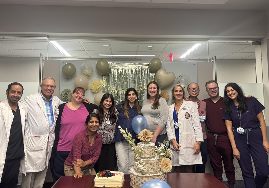 A group of healthcare team members gather around a table with cakes and baby shower decorations in an office or conference room. Balloons, floral decorations, and a decorative backdrop are displayed behind the group.