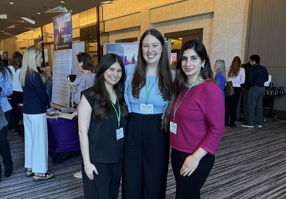 Three conference attendees wearing name badges pose together in an event venue. Other attendees, informational displays, and vendor tables are visible in the background.